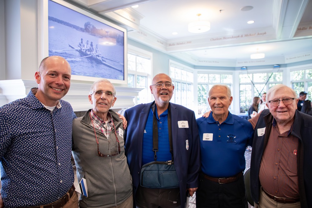 Group of alumni pose together in front of tv displaying a picture of rowing